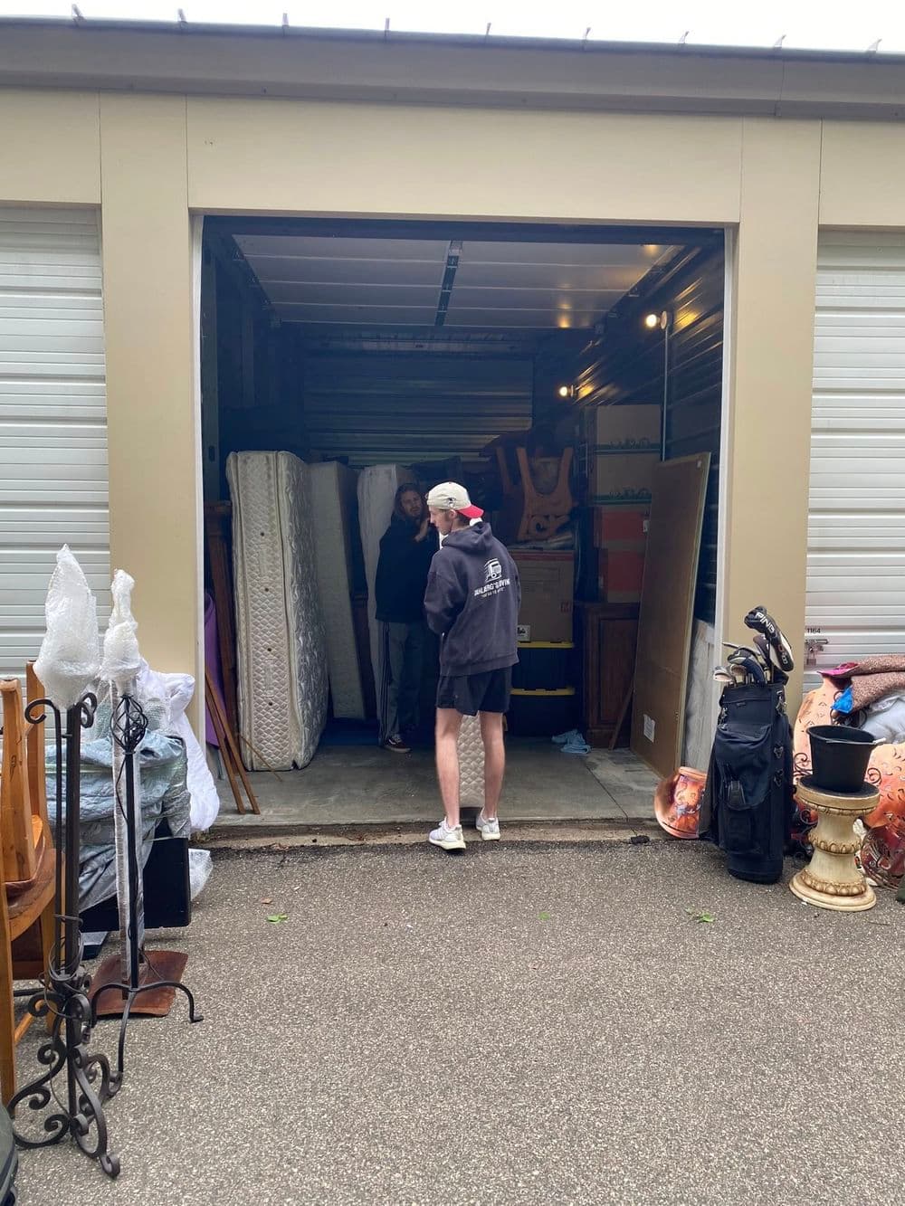 Person standing in a storage unit filled with furniture, mattresses, and various household items.