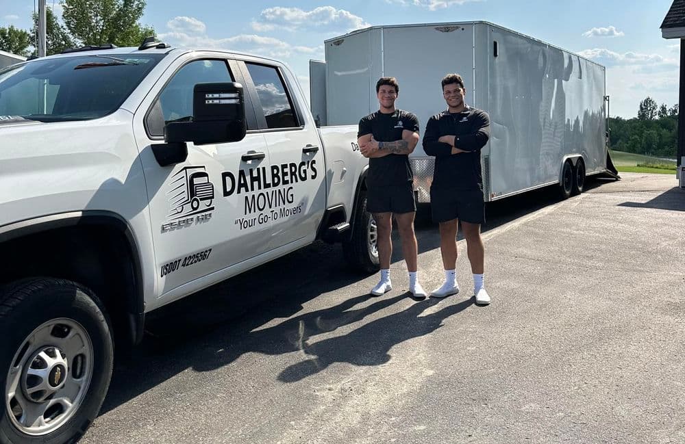 Young movers standing by a Dahlberg's Moving truck with a trailer, ready to assist customers.
