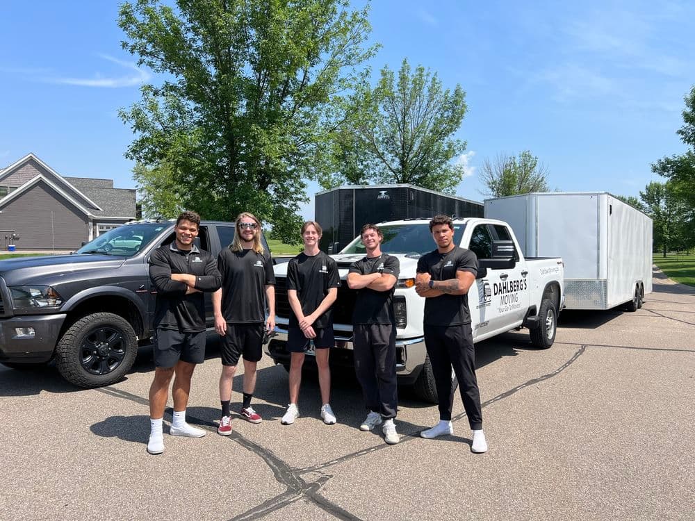 Group of five movers posing confidently in front of a truck and trailer in a residential area.